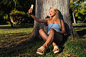 Unbeschwerte Frau, die sich mit ihrem Smartphone selbst porträtiert und Musik über Kopfhörer hört, während sie unter einem Baum im Park bei Sonnenuntergang im Sommer sitzt