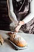Anonyme Handwerkerin in Schürze bestreut einen köstlichen Laib Weißbrot mit Mehl am Tisch in einer Backstube