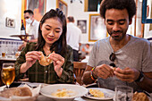 Happy young multiethnic couple in casual clothes smiling while eating delicious foods during date in modern restaurant