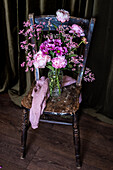 Strauß frischer bunter Pfingstrosen und Chrysanthemen in einer Glasvase auf einem verwitterten Holzstuhl neben Vorhängen in einem hellen Raum