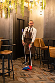 Full body of busy male waiter with scoop and broom cleaning floor in modern restaurant during coronavirus pandemic looking at camera