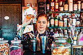 Female barkeeper pouring alcohol in shaker while preparing refreshing cocktail at counter in bar and looking away