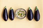 Top view of fresh ripe aubergines placed on beige table with bowl of appetizing traditional Baba ghanoush dish