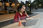 Junge Bloggerin sitzt mit gesundem Essen am Tisch, benutzt Laptop und telefoniert, während sie in einem modernen Restaurant Zeit verbringt