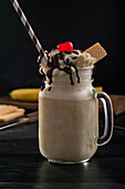 Glass jar of sweet banana split milkshake topped with whipped cream waffles chocolate and cherry on table