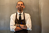 Male waiter with tattoos in white shirt and apron looking at camera while standing on gray background during work in modern cafe