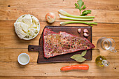 Top view of raw marinated beef brisket for corned placed on chopping board with vegetables and spices on wooden table