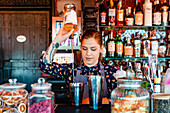 Female barkeeper pouring alcohol in shaker while preparing refreshing cocktail at counter in bar and looking away