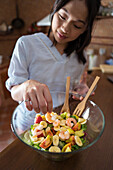 Asian female putting fresh shrimps on top of salad in glass bowl in kitchen at home