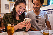 Young Asian female showing photos on smartphone to ethnic boyfriend sitting at table in restaurant