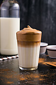Ein Glas köstlicher Dalgona-Kaffee mit Milch und schaumigem Topping steht auf einem schwarzen, unordentlichen Tisch mit Kakaopulver und Zucker