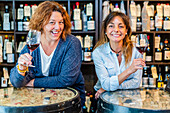 Lächelnde Freundinnen in legerer Kleidung, die einen aromatischen Rotwein probieren und in die Kamera schauen, vor einem Regal mit Weinflaschen in einem Restaurant