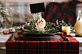 Weihnachtlich gedeckter Tisch mit Rentier aus Holz mit leerer Namenskarte auf Keramikteller auf rot-kariertem Tischtuch im Hintergrund