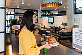 Side view of content female with tasty burger on plate looking forward against signboard in restaurant