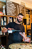 Focused man sommelier with beard and moustache in black apron standing at bar counter and pouring bottle of red wine into glass decanter in restaurant