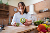 Soft focus of female demonstrating half of fresh kiwi over chopping board while cooking salad for lunch at home