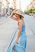 Seitenansicht einer fröhlichen Frau mit Strohhut und Sommerkleidung, die auf der Straße steht und den Sommertag in der Stadt genießt