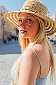 Seitenansicht einer charmanten Frau mit Strohhut, die an einem sonnigen Tag auf einer Straße in der Stadt in die Kamera schaut