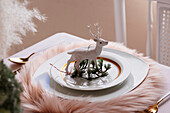 From above christmas table settings with a wreath decoration and white and golden dinnerware and white reindeer on fluffy placemat on pink tablecloth