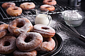Sweet fried doughnuts served on plate near metal cooling rack and jug of milk on black messy table with powdered sugar
