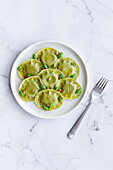 Top view of tasty raviolis with basil decorated with pine nuts served on white round plate on table in light kitchen