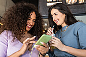 Lächelnde junge Freundinnen in Freizeitkleidung, die bei einer Limonade im Restaurant auf ihrem Handy surfen