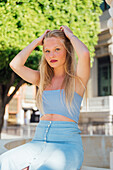 Blonde female in summer outfit sitting in street on sunny day and looking at camera while enjoying summer