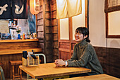 Zufriedene asiatische Frau in legerem Pullover, die mit einem breiten Grinsen in die Kamera schaut, während sie an einem Holztisch in einer Ramen-Bar sitzt