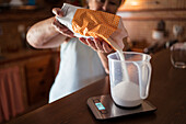 Ältere Frau gießt Zucker aus Papiertüte in Krug auf Waage beim Kochen in Küche