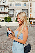 Schöne blonde junge Frau isst kaltes leckeres Eis, während sie an einem sonnigen Sommertag auf der Straße steht