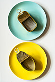 Draufsicht auf rechteckige und ovale geöffnete leere Metalldosen, die auf gelben und blauen Tellern auf einem weißen Tisch stehen