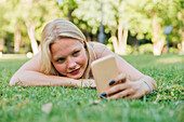 Lächelnde, charmante Frau, die im Park im Gras liegt, ein Selfie mit ihrem Smartphone macht und mit Kopfhörern im Sommer Musik hört