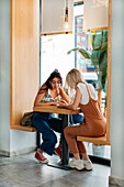 Best female friends surfing internet on cellphone while talking at cafeteria table in daylight