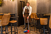 Beschäftigter männlicher Kellner mit Schaufel und Besen beim Reinigen des Fußbodens in einem modernen Restaurant während einer Coronavirus-Pandemie