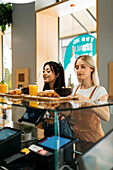 Side view of young cheerful best friends receiving trays with delicious food and beverages from crop anonymous vendor in cafeteria