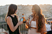 Seitenansicht positiver junger multiethnischer Freundinnen, die fröhlich lächeln und mit Bierflaschen anstoßen, während sie bei Sonnenuntergang auf einer Terrassenbar in Kappadokien, Türkei, eine angenehme Zeit miteinander verbringen