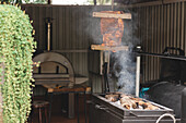 Leckere Schweinerippchen hängen über einem modernen heißen Grill mit Feuerholz und Rauch während des Kochvorgangs auf der Terrasse eines hellen Cafés