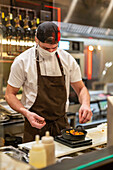 Through glass of male cook in apron and cap with protective mask sprinkling condiment on top of meal in pan in restaurant during coronavirus pandemic