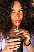 Crop glad young female with long curly hair sipping cold fizzy drink with straw and looking away coquettishly