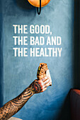 Crop unrecognizable person showing tasty sandwich to go against wall with The Good The Bad And The Healthy inscription