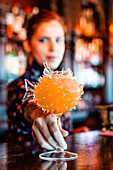 Unscharfe Barkeeperin mit alkoholischem Orangencocktail in einem fischförmigen Glas auf dem Tresen einer Bar