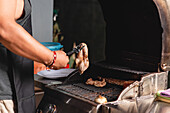 Cropped unrecognizable male in apron placing raw marinated meat on grill of barbecue cooker while cooking steaks