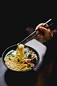 Hand of unrecognizable woman picking noodles with chopsticks from bowl of savory Asian soup in cafe