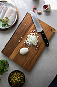 From above wooden board with cut onion and knife placed near peas and hake fillet with herbs during food preparation in kitchen