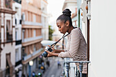 Seitenansicht einer jungen Afroamerikanerin mit professioneller Fotokamera auf einem umzäunten Balkon in der Stadt