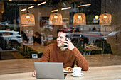 Through window of happy young male student sitting at table with laptop and cup of coffee with croissant and talking on mobile phone while spending time in modern cafeteria