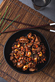 Schale mit asiatischem Hähnchenfleisch mit Mandeln und Gemüse in der Nähe von Bambus-Essstäbchen und Schnittlauch von oben