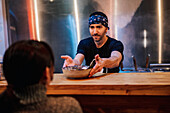 Bearded male in bandana and black t shirt putting plate with noodle on wooden counter for client in ramen bar