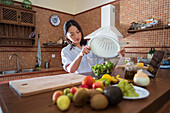 Young Asian female putting lettuce leaves from colander into glass bowl while cooking healthy salad for lunch in kitchen at home