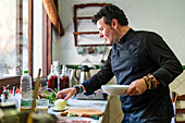 Seitenansicht eines gutaussehenden männlichen Kochs in schwarzer Uniform, der Teller hält, während er eine Mahlzeit zubereitet und neben einem Tisch mit verschiedenen Zutaten in der Küche steht
